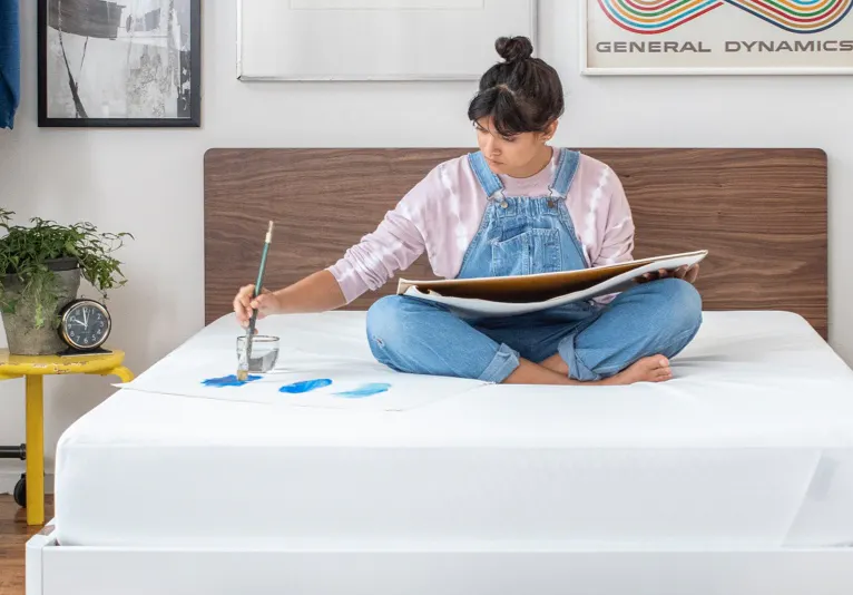A woman paints while sitting on a mattress.