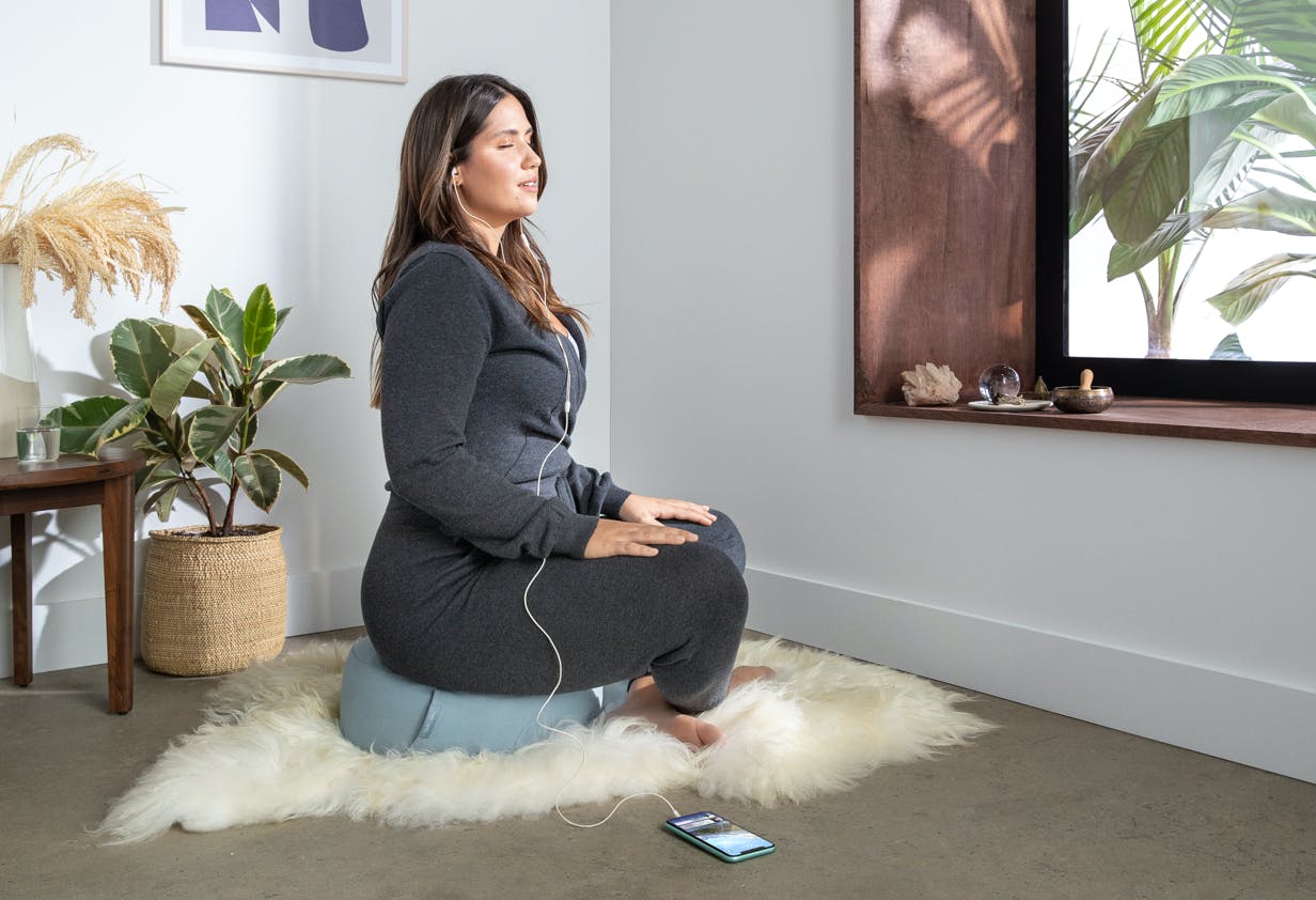 A woman sits on a light blue meditation cushion with headphones on.