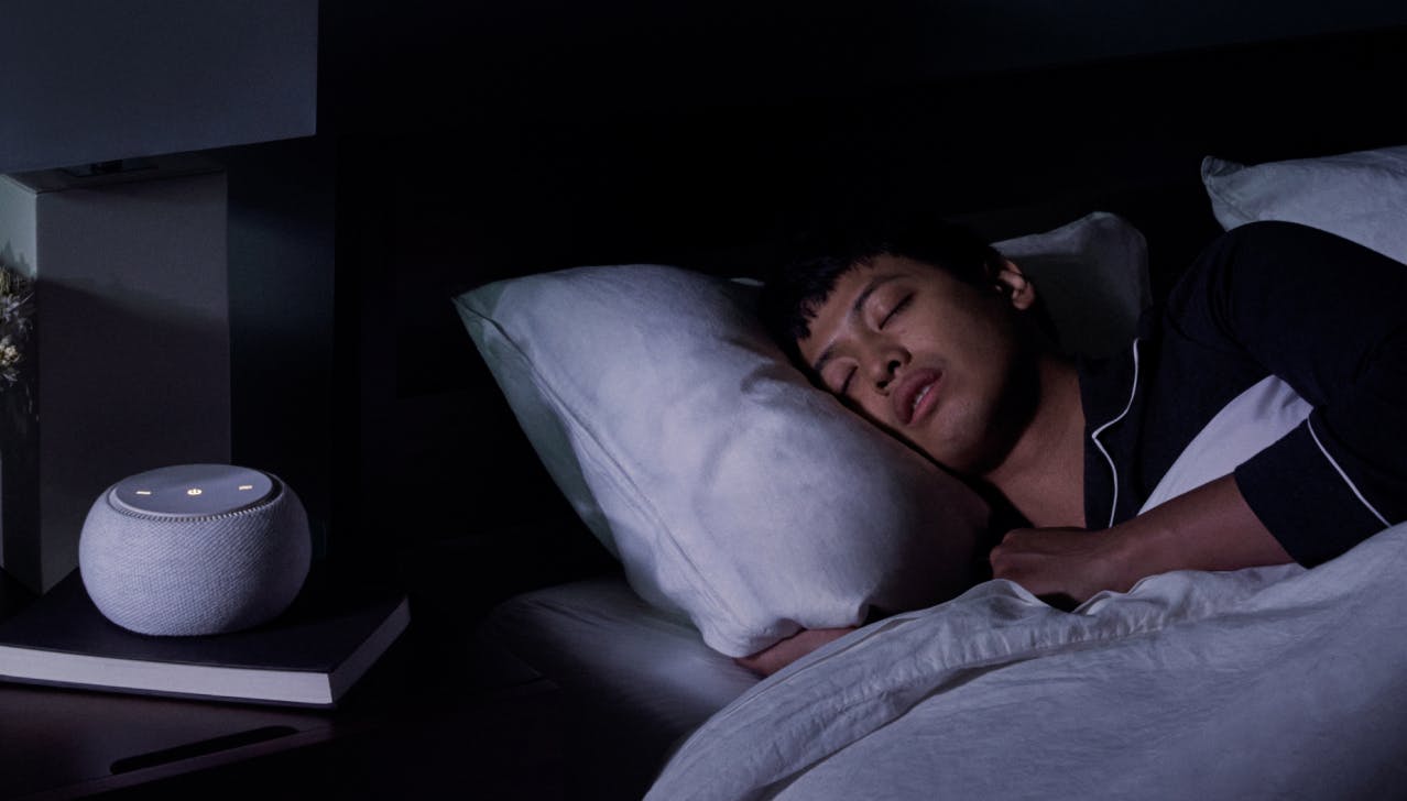 Man sleeping in bed with White Noise Machine on nightstand.