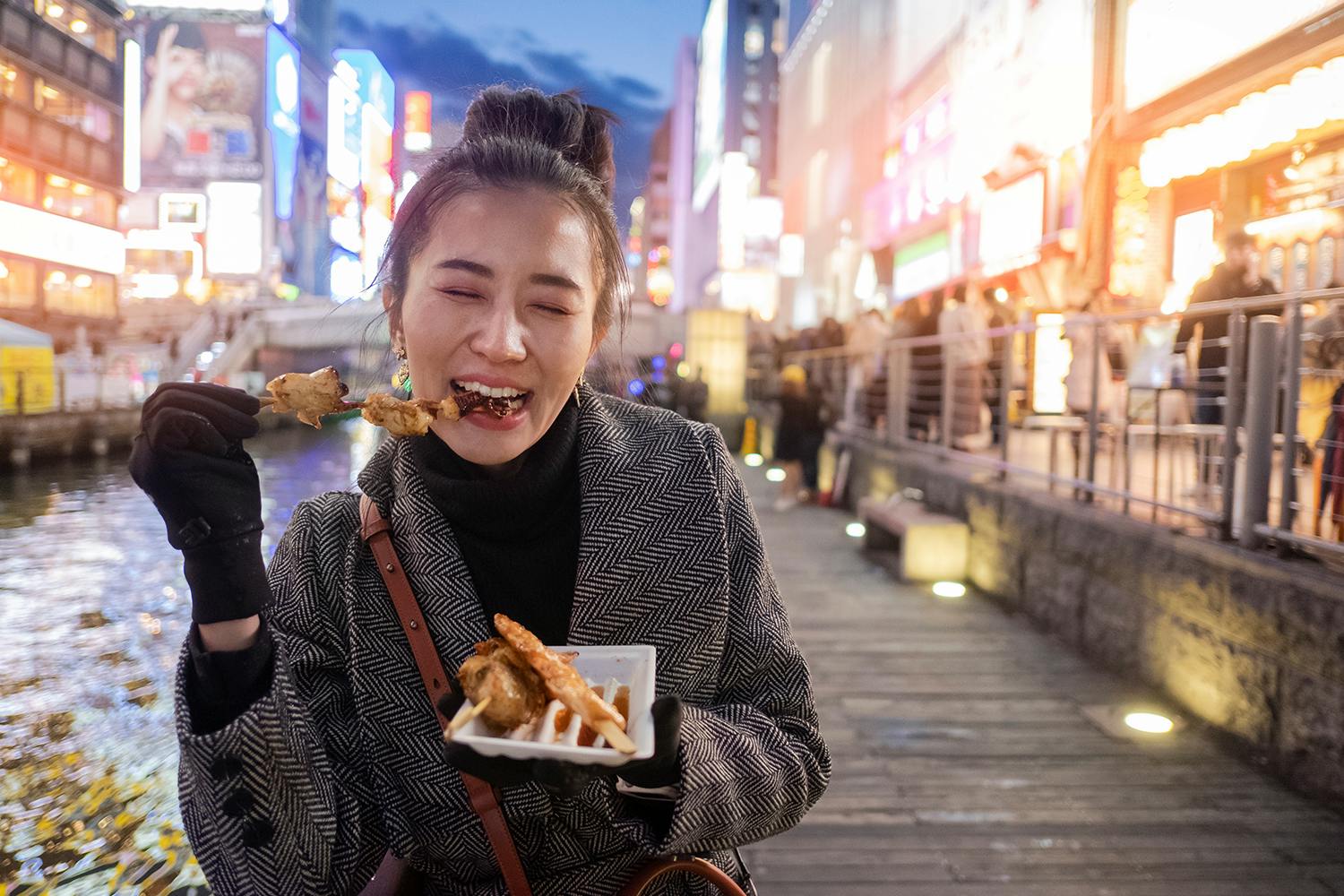 A woman is eating street food in Osaka near Dotonbori street with neon Japanese signs in the background