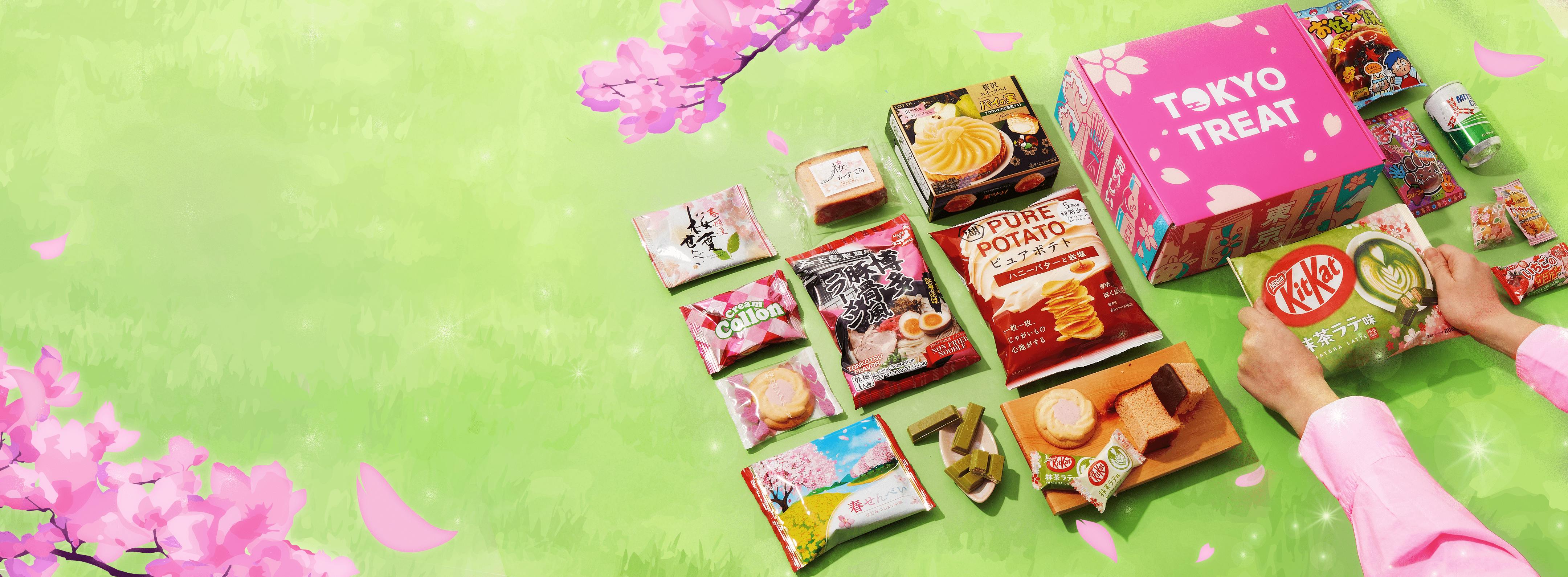 The TokyoTreat special-edition Sakura box sits against a cherry blossom backdrop surrounded by green grass and sakura snack items.