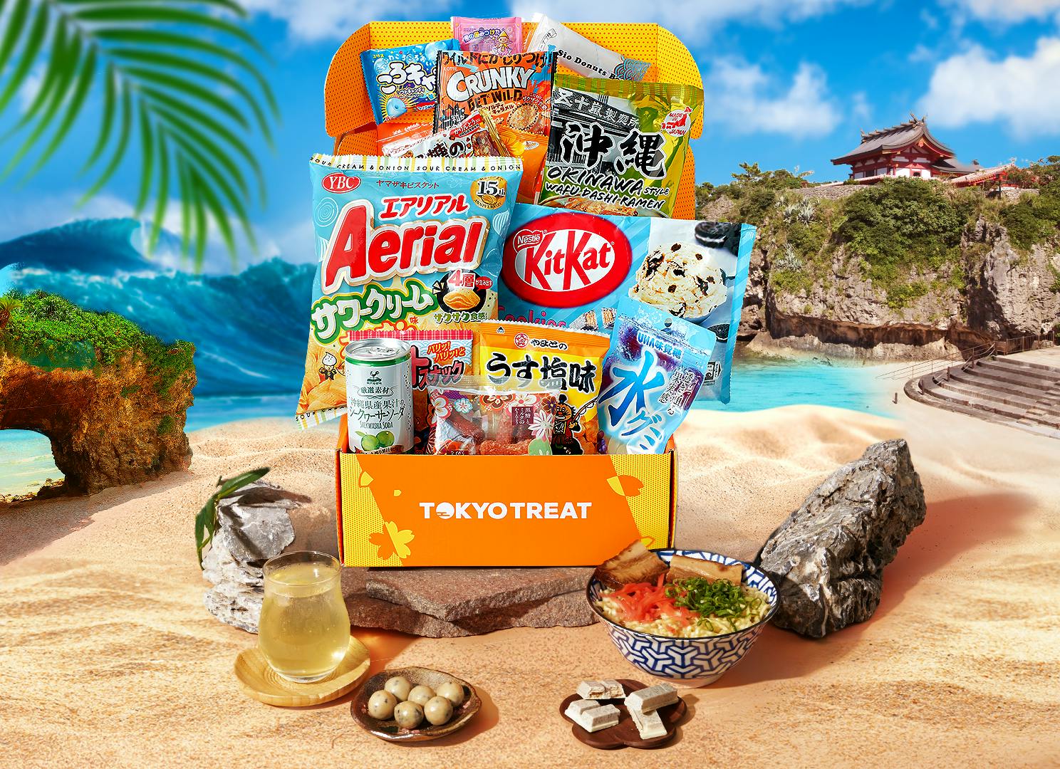 The Okinawa Snackin' Oasis box sits against a backdrop of a beach in Okinawa, with waves and an Okinawa castle in the background.