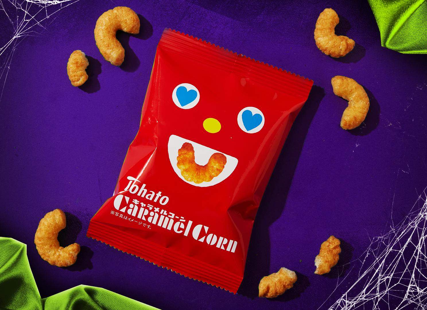 The bright red bag of Tohato Caramel Corn Puffs sits against a purple backdrop, with green fabric and spider webs at the corners.