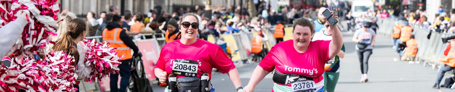 Two women running hand in hand wearing a pink Tommy's running top, being cheered on by cheerleaders