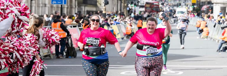 Two women running hand in hand wearing a pink Tommy's running top, being cheered on by cheerleaders