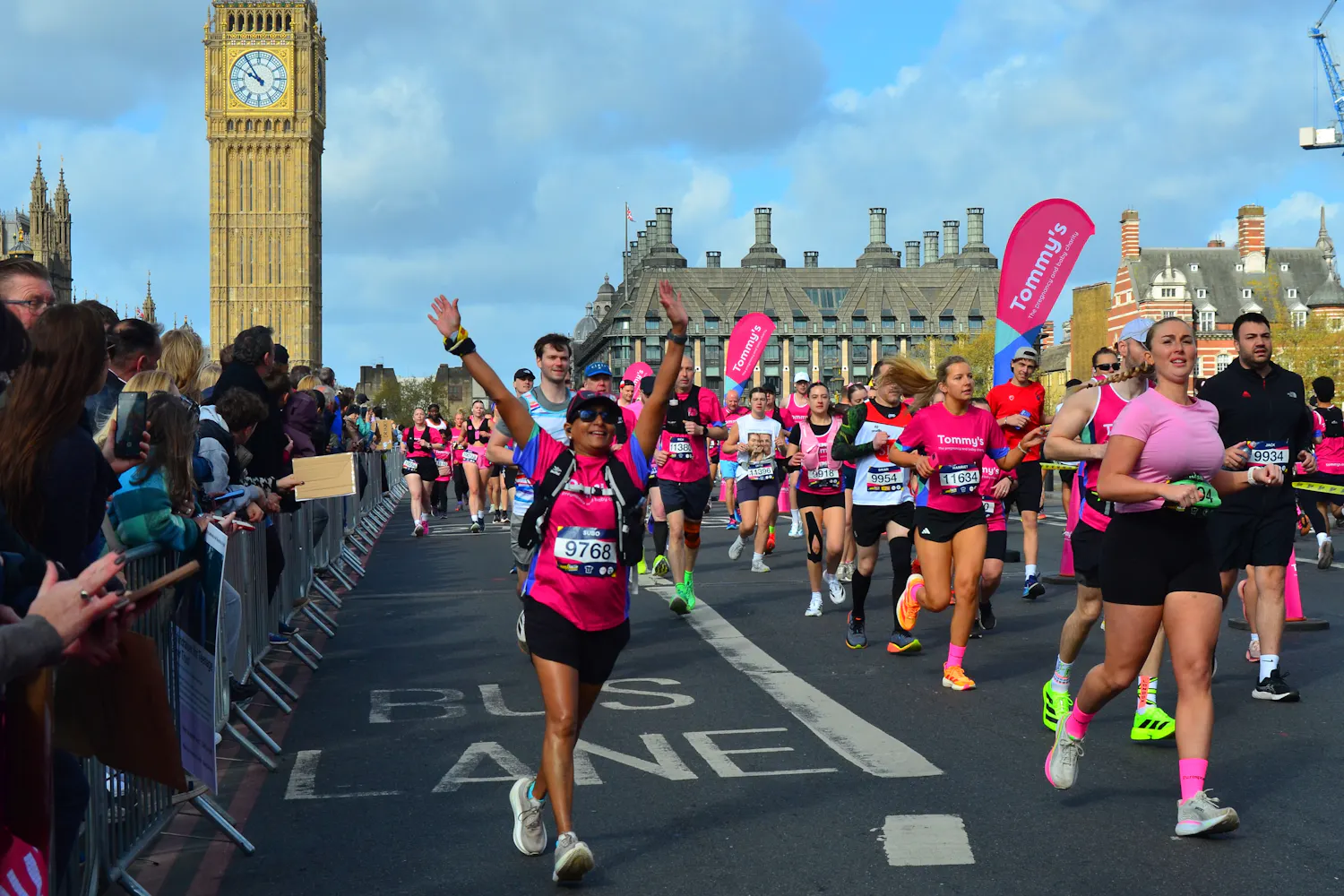 Runner in pink Tommy’s shirt raises arms while passing Big Ben and cheering spectators.