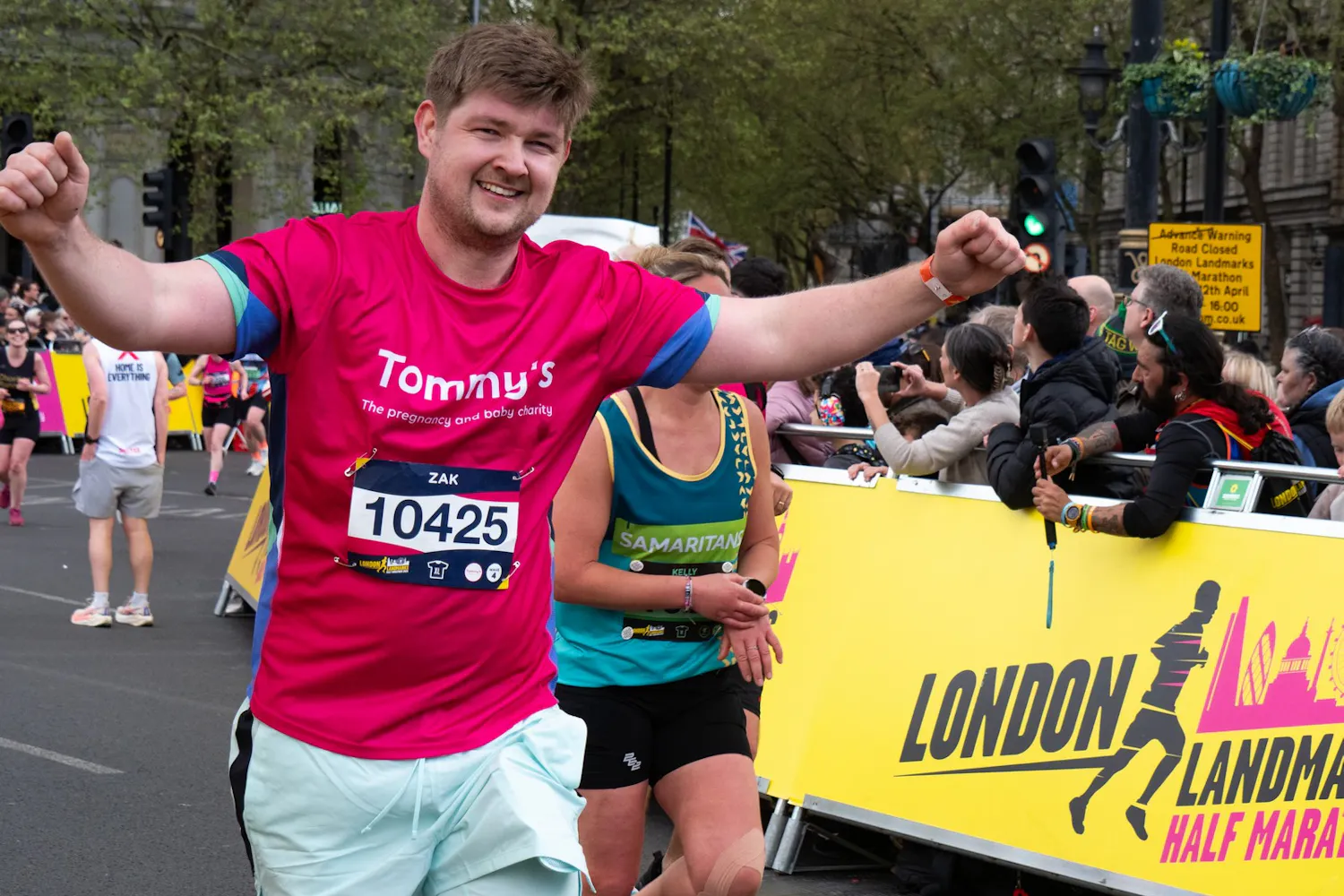 Runner in pink Tommy’s shirt spreads arms near London Landmarks Half Marathon signage.