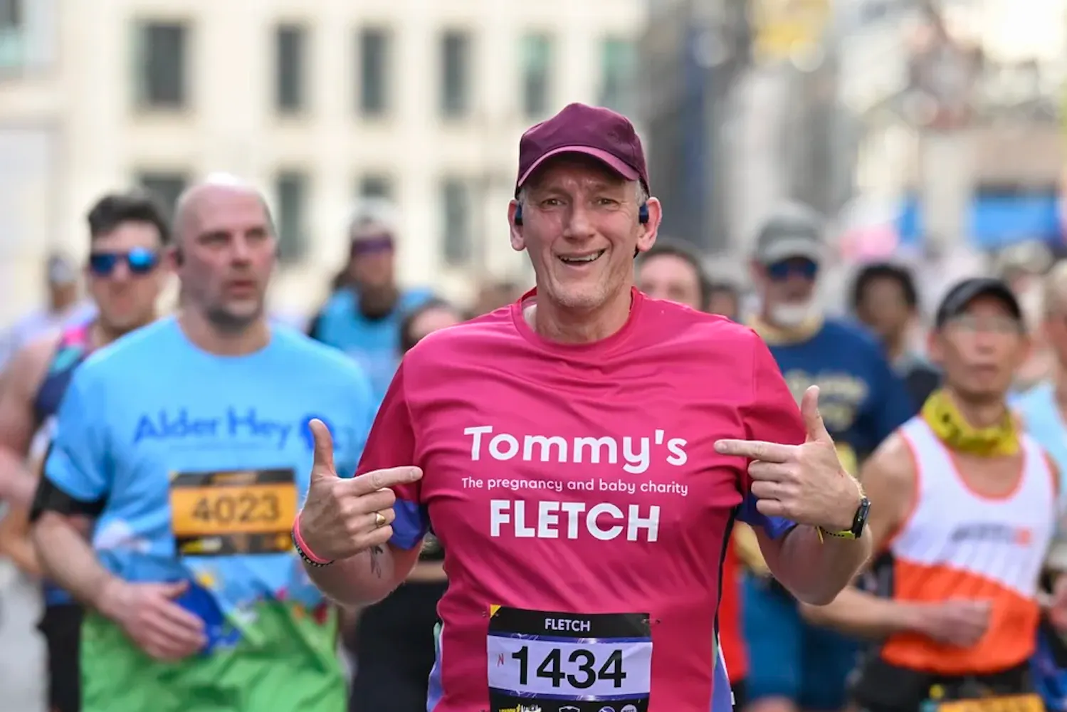 Runner in pink Tommy’s top points to charity logo while running among marathon participants.