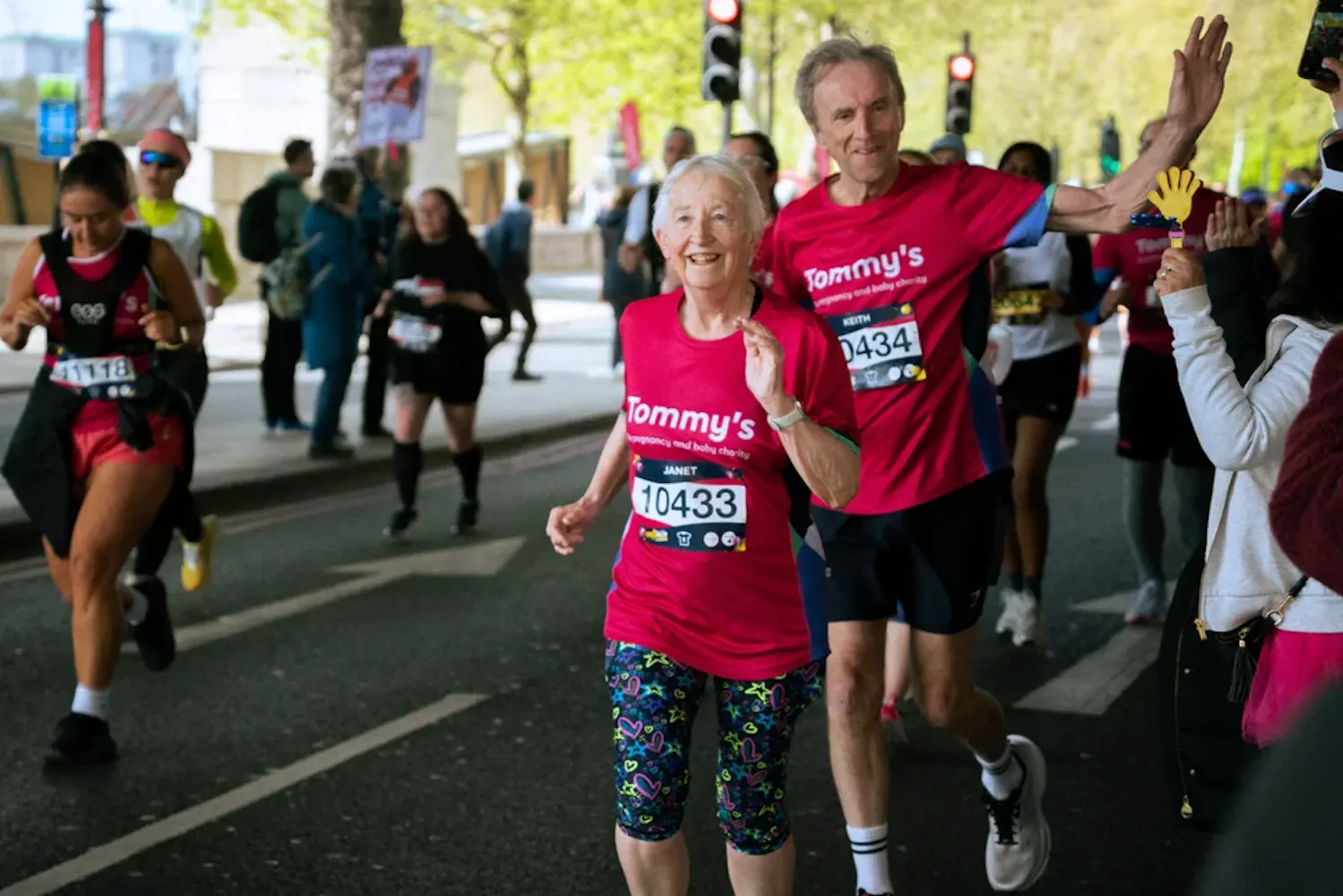 Two runners in pink Tommy’s shirts wave while running past spectators along city marathon route.