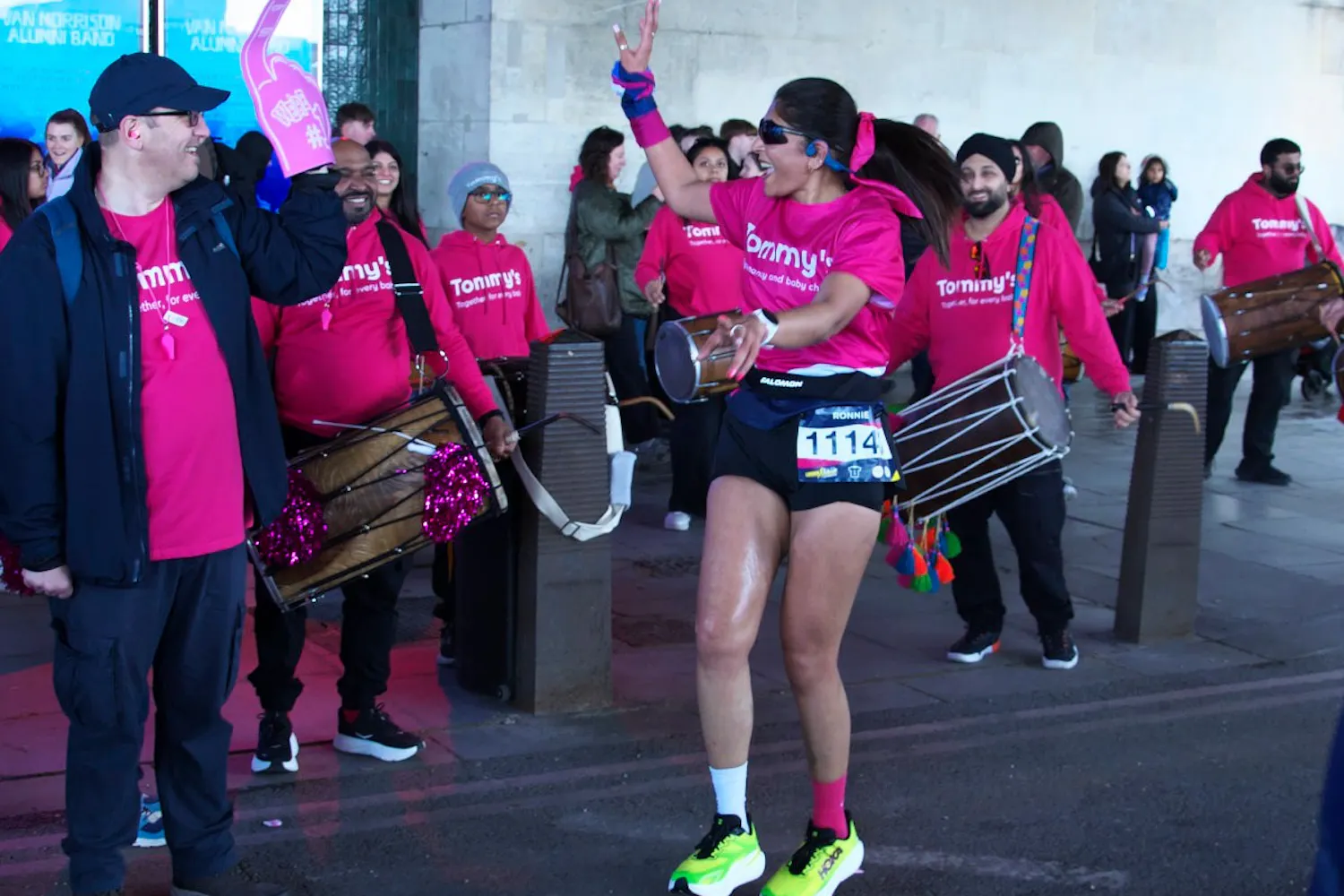 Runner in pink Tommy’s top dances alongside drummers cheering on marathon route.