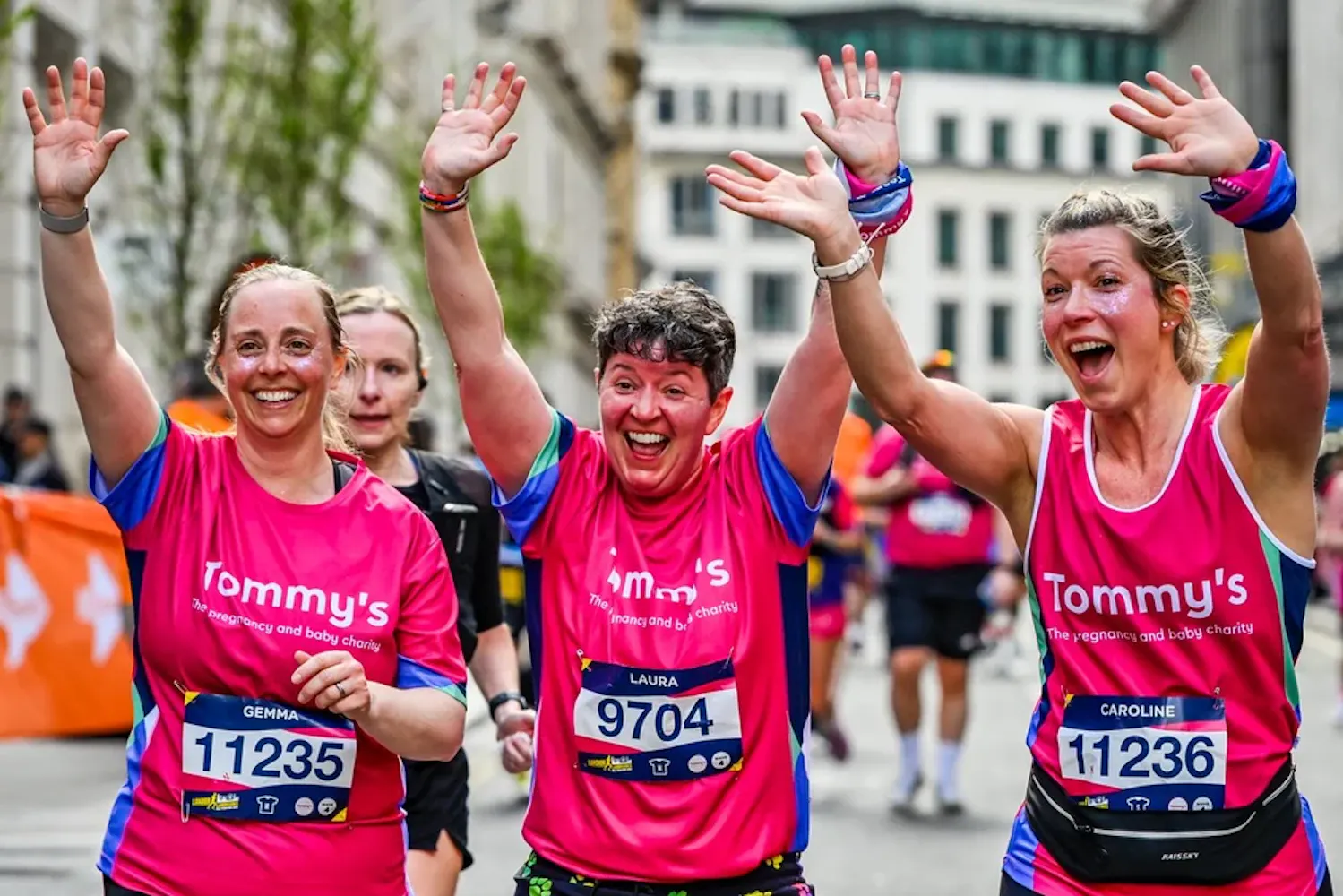 Three runners in pink Tommy’s tops raise hands and smile while running together during race.