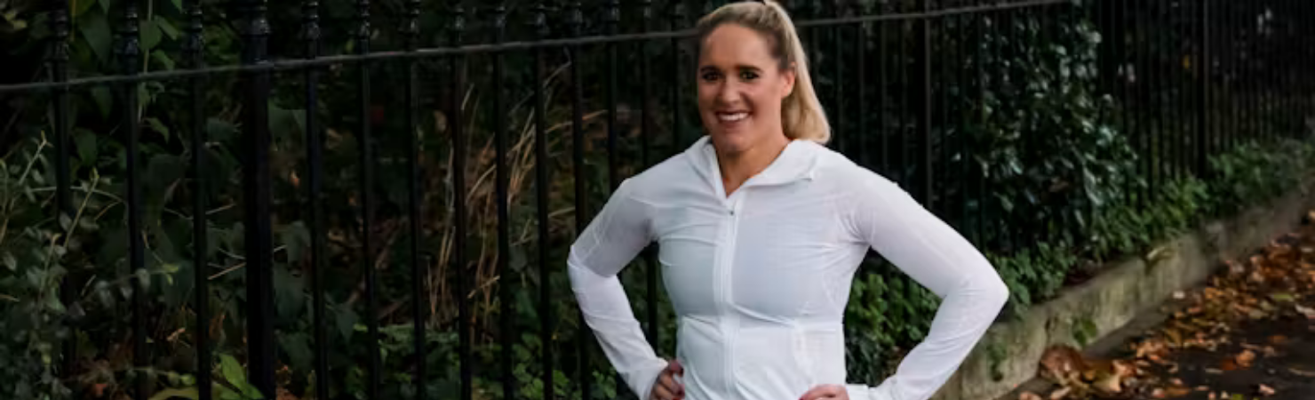 Person doing a lunge stretch on a pavement beside a metal fence, wearing a white long-sleeve top, black leggings and trainers, with fallen leaves on the ground.