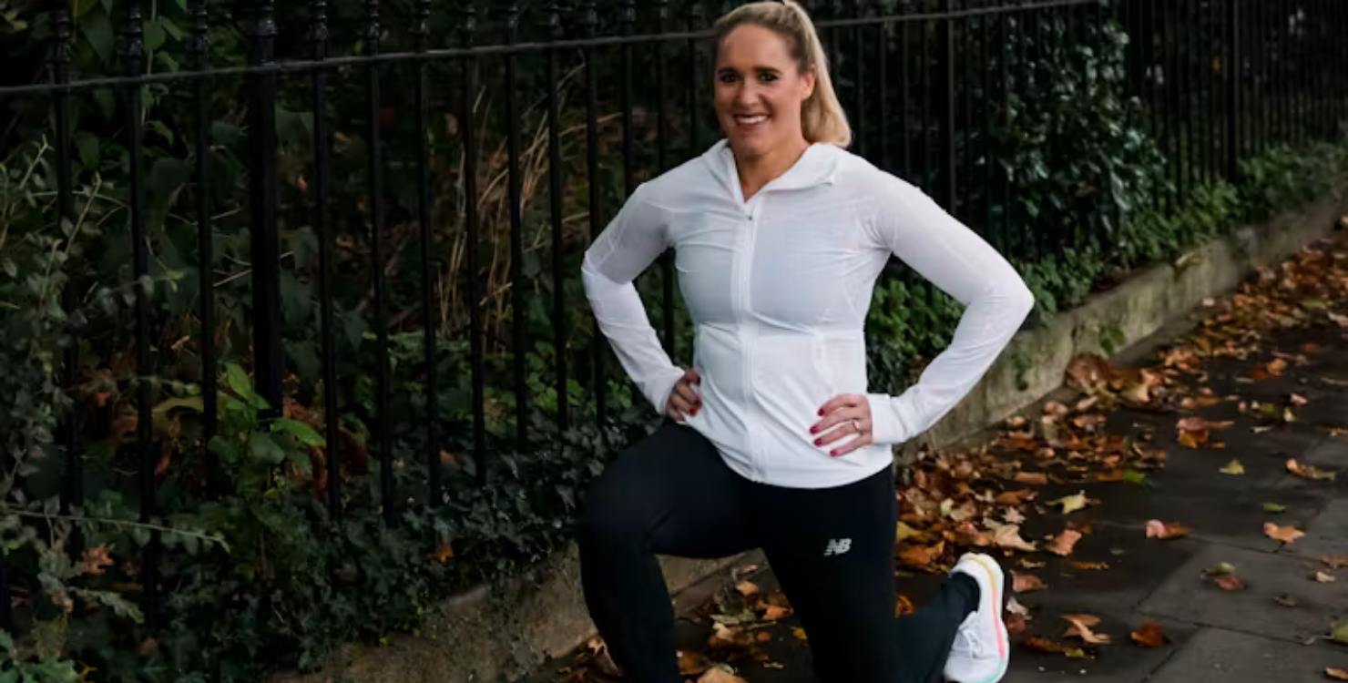 Person doing a lunge stretch on a pavement beside a metal fence, wearing a white long-sleeve top, black leggings and trainers, with fallen leaves on the ground.