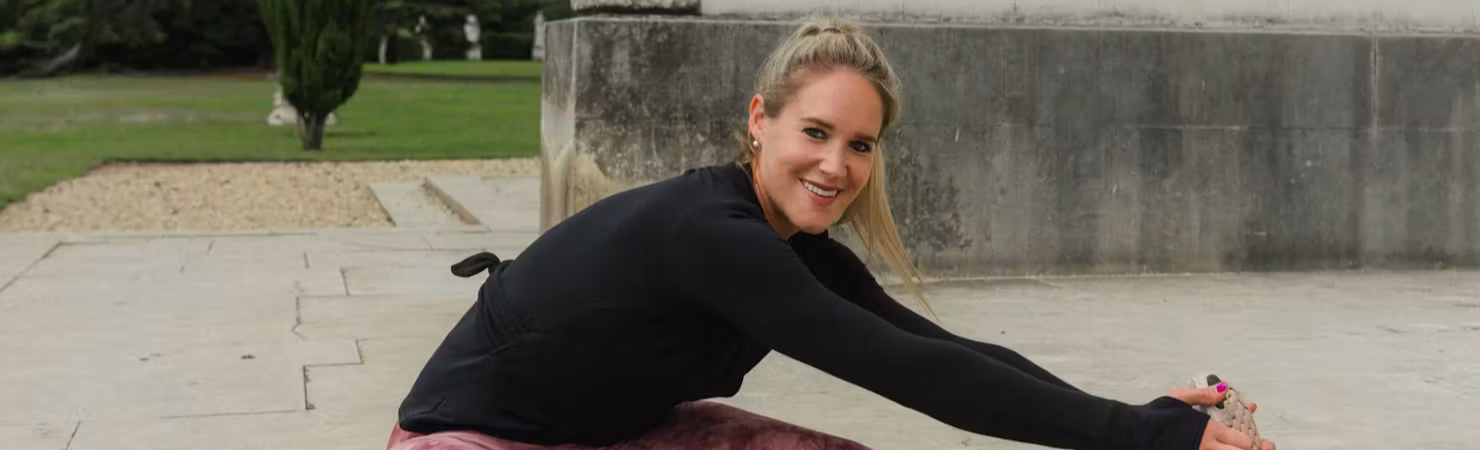 Person sitting on a mat outdoors, stretching one leg forward and holding their foot, wearing a black top, patterned leggings and trainers.