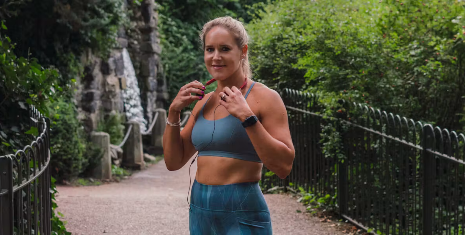 Person standing on a park path adjusting earphones, wearing a sports bra and leggings, with greenery and a stone wall in the background.