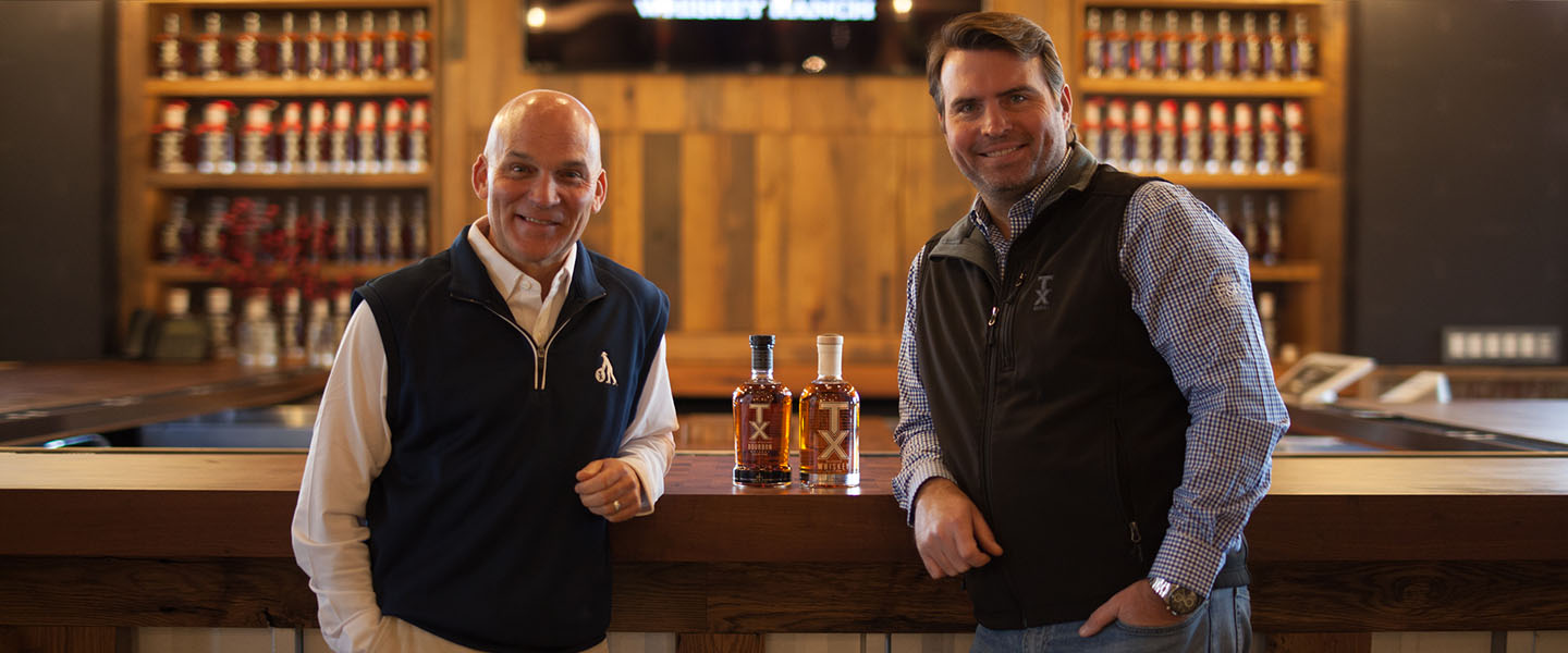 Leonard Firestone and Troy Robertson founders of Firestone and Robertson Distilling Co. standing next to two bottles of TX Whiskey.