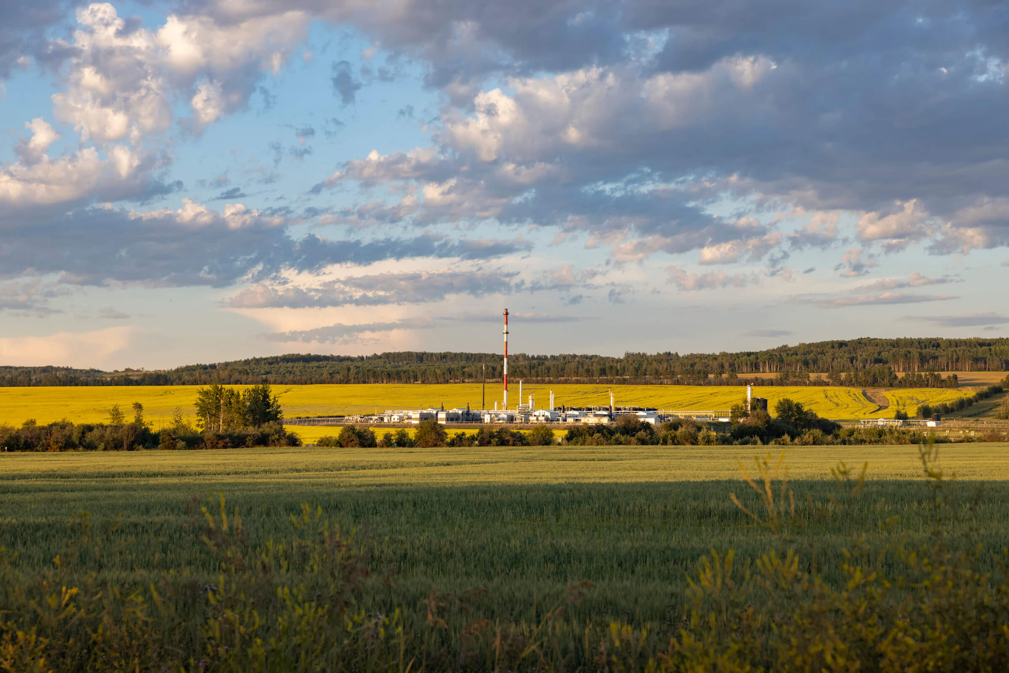 A gas plant in a green field under a cloudy blue sky.