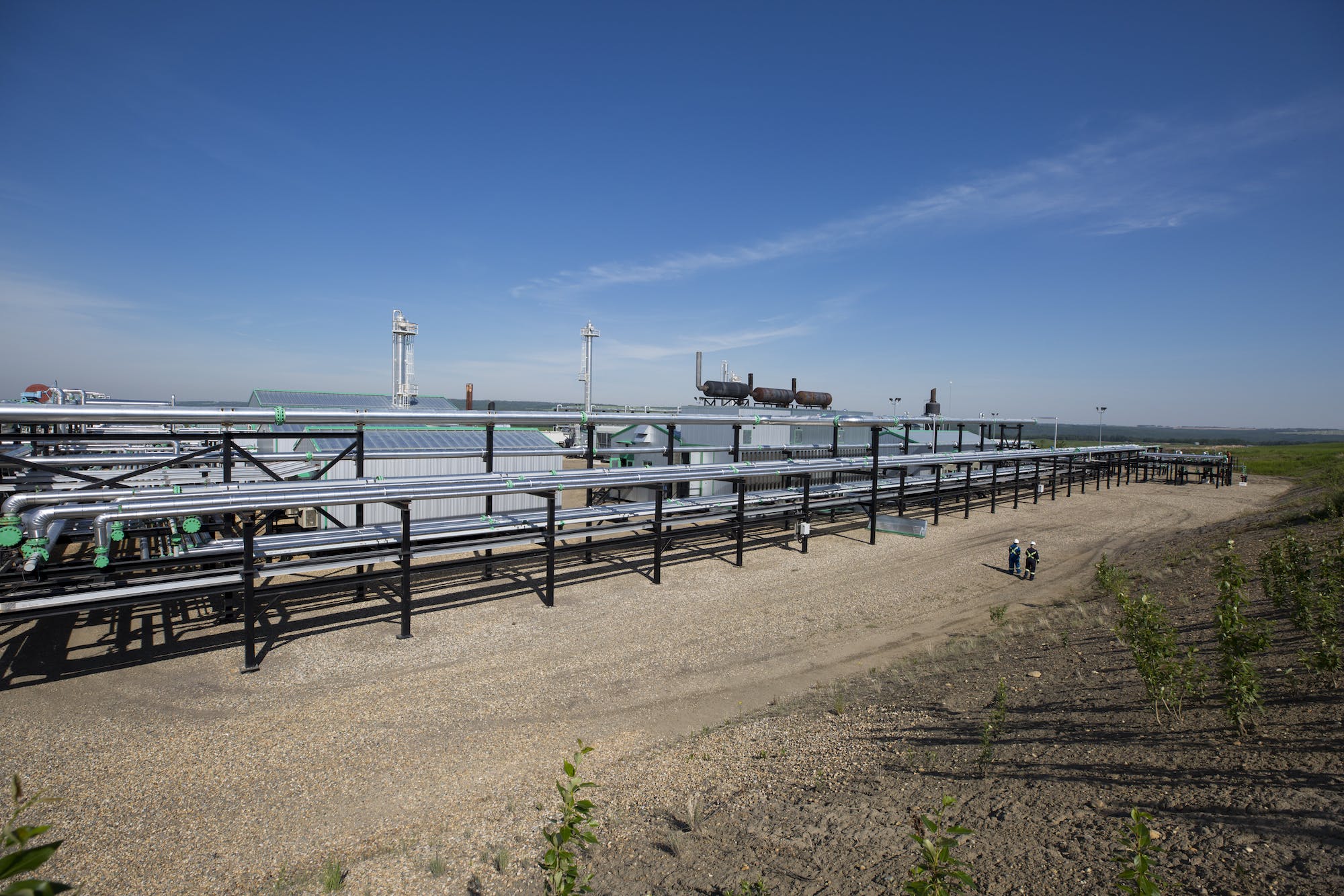 A wide view of long silver pipes at a gas plant, and a few workers standing in the distance.