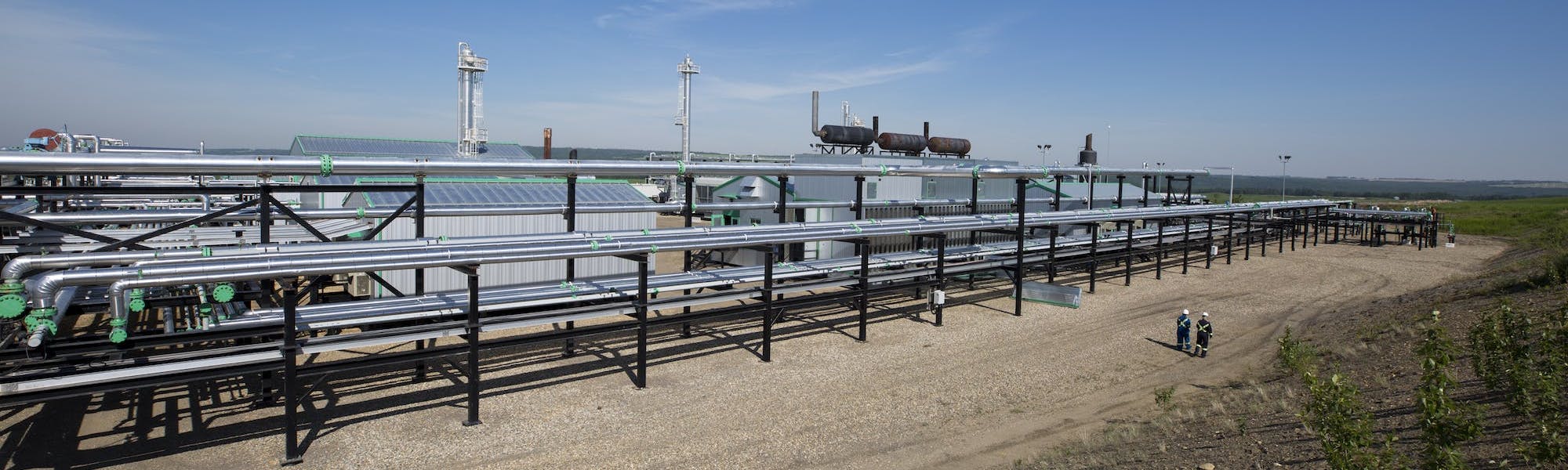 A wide view of long silver pipes at a gas plant, and a few workers standing in the distance.