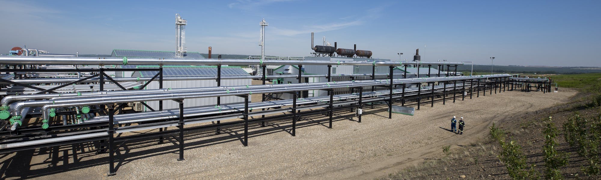 A wide view of long silver pipes at a gas plant, and a few workers standing in the distance.