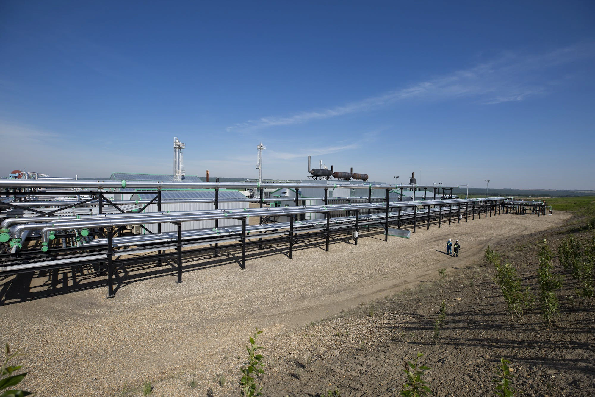 A wide view of long silver pipes at a gas plant, and a few workers standing in the distance.