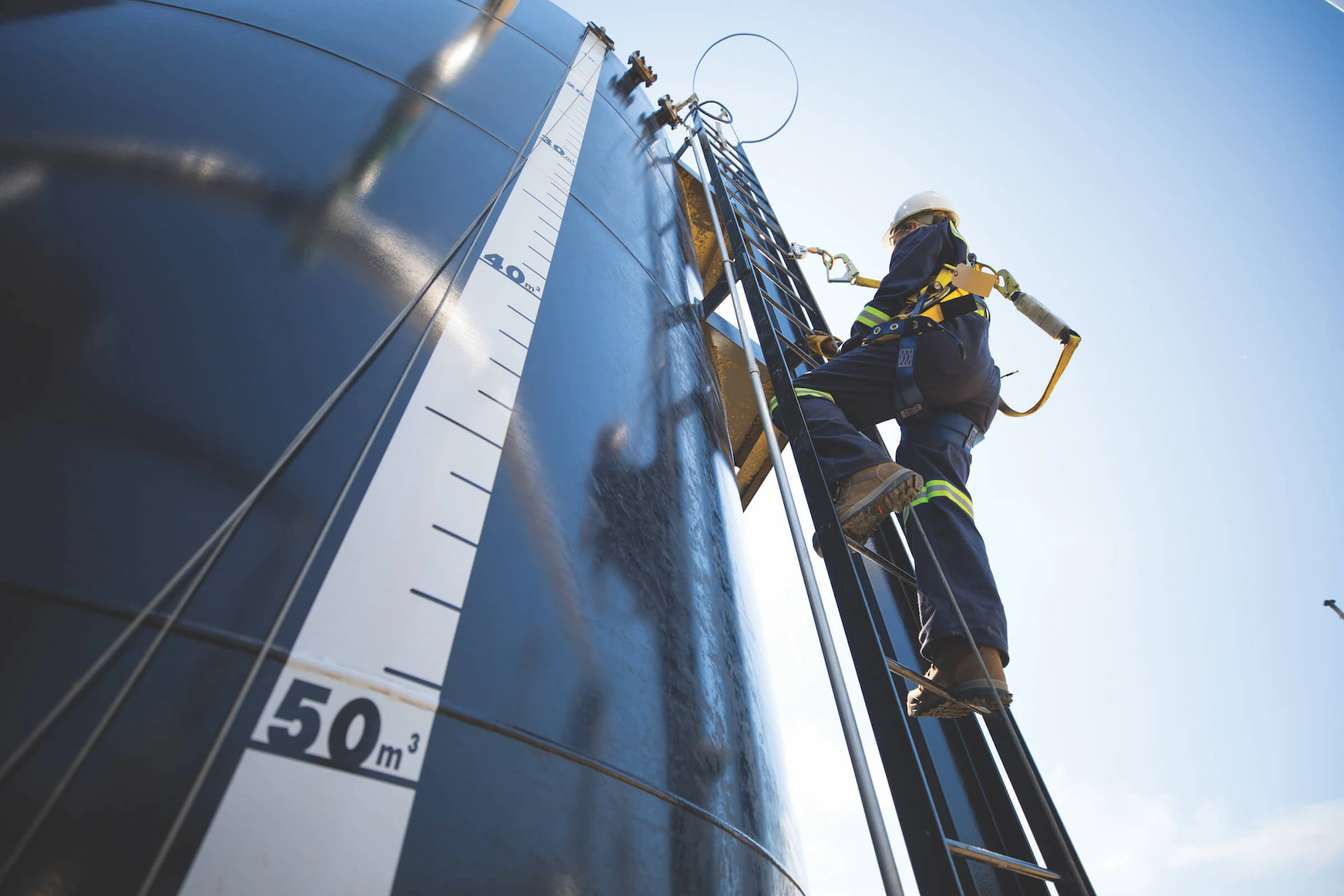 A worker in blue coveralls and a white hard-hat scales a ladder attached to a tank with measurement values printed on the side.
