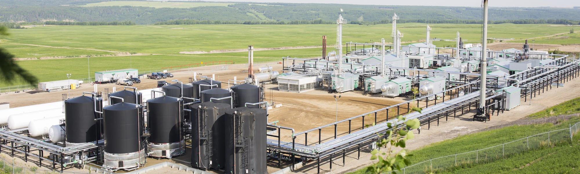 A hilltop view of a gas plant in a green field, with a few pine branches in the foreground.