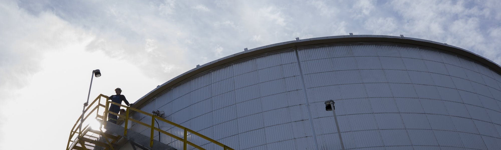 An oil rig worker standing at the top of a yellow staircase on a large tank.