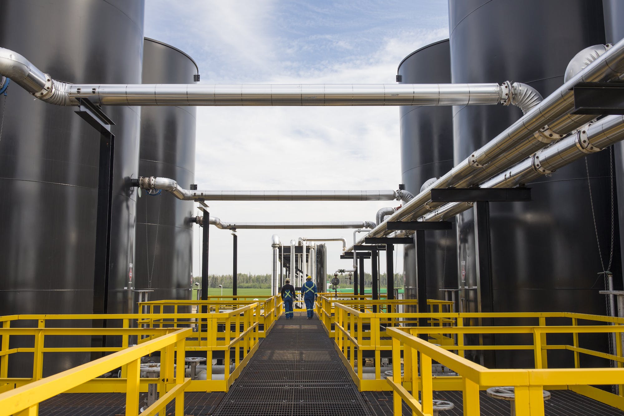 Two gas plant workers walk between large tanks on a walkway with yellow railings.