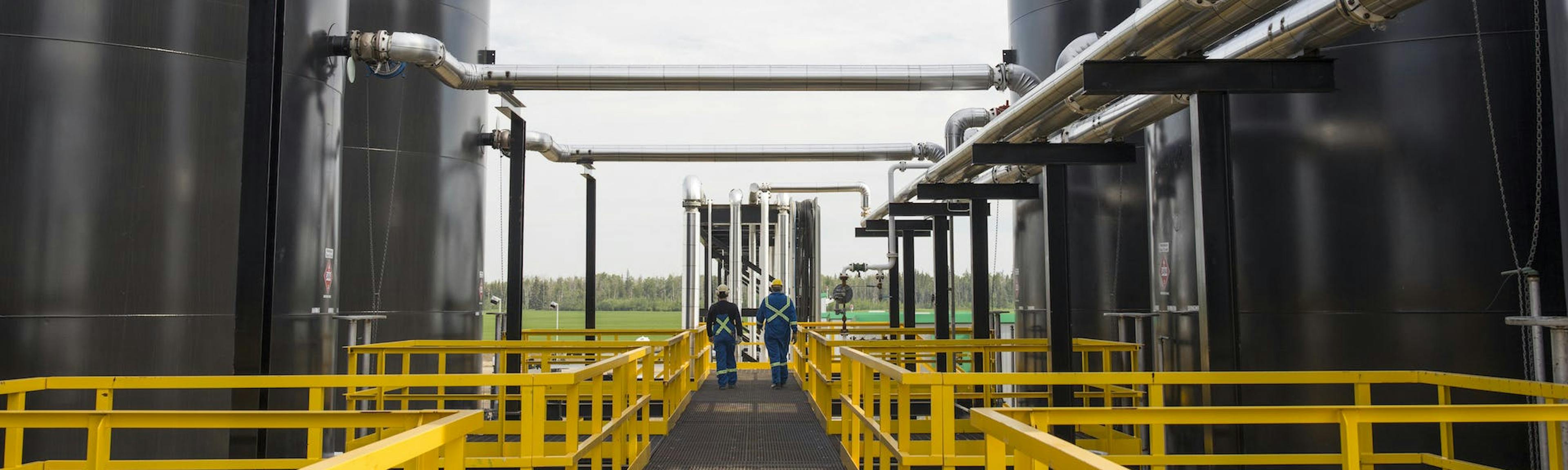 Two gas plant workers walk between large tanks on a walkway with yellow railings.