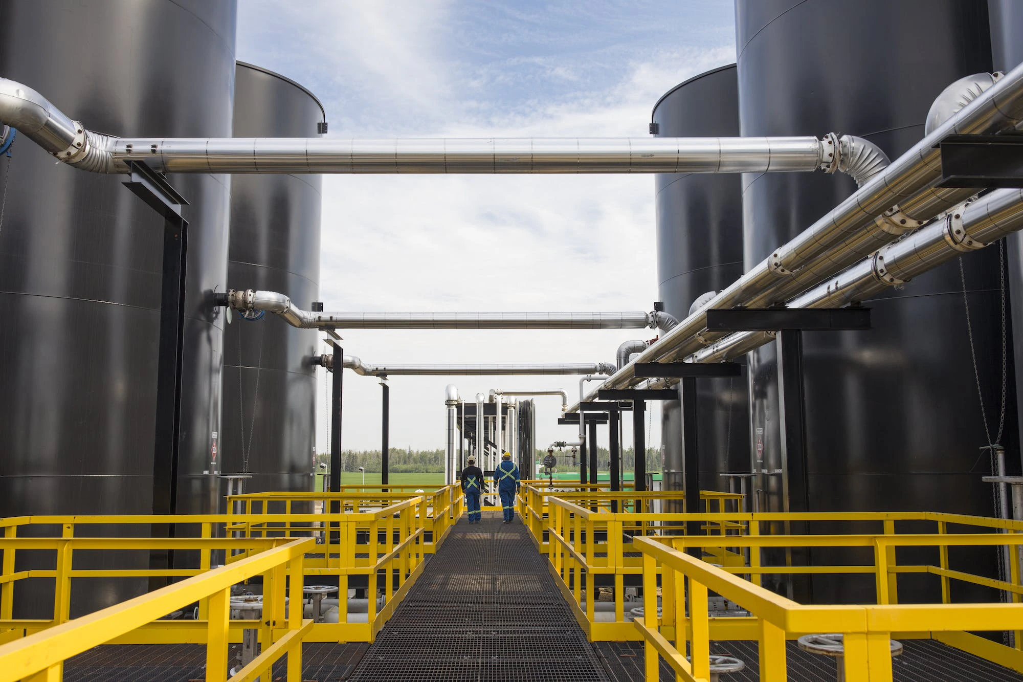 Two gas plant workers walk between large tanks on a walkway with yellow railings.