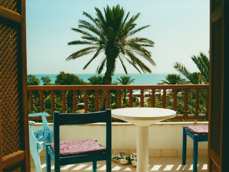 A sweet balcony scene with modernist furniture overlooking a beach with palm trees.