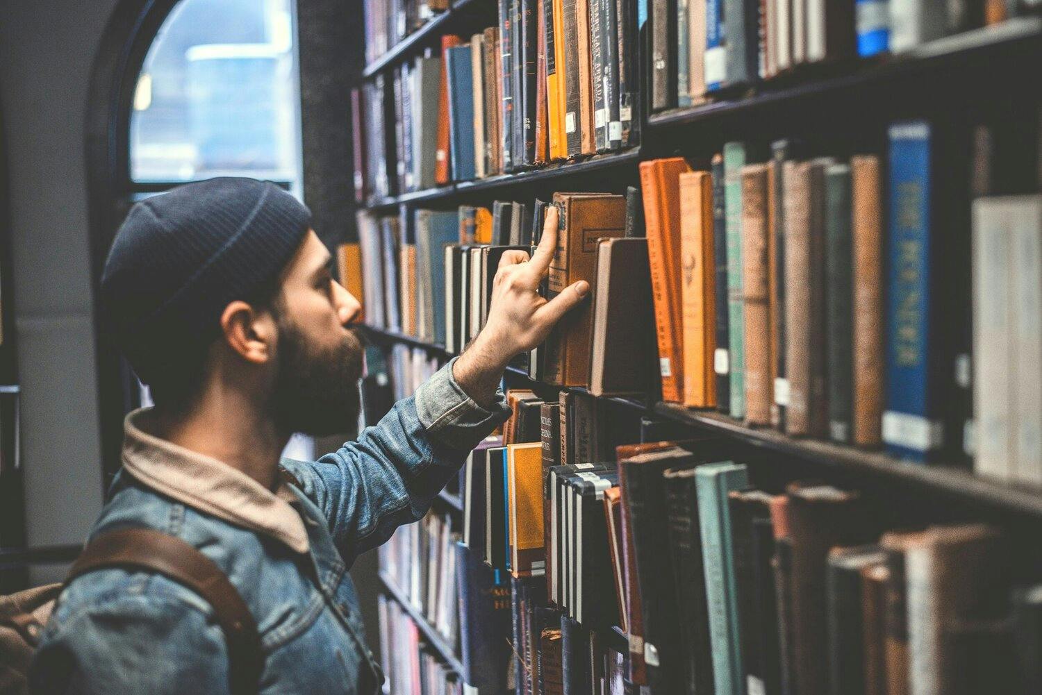Bearded man in hat selects a book from a library shelf.