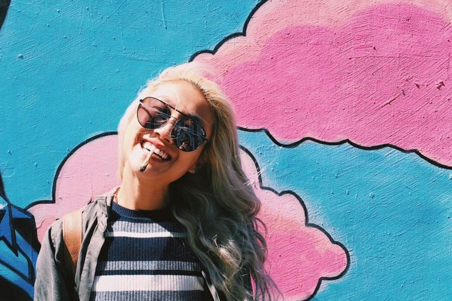 Young woman smiling and wearing sunglasses, holding a joint between her lips and standing in front of a wall painted sky blue with pink clouds.