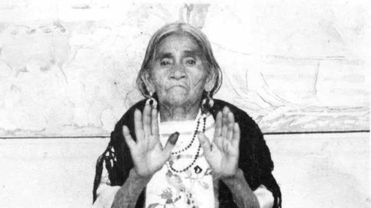 María Sabina, holding up her hands while standing against a wall, replete with beads around her neck. 