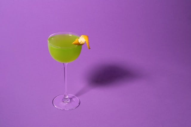 A green cocktail with an orange rind twist against a purple backdrop. 