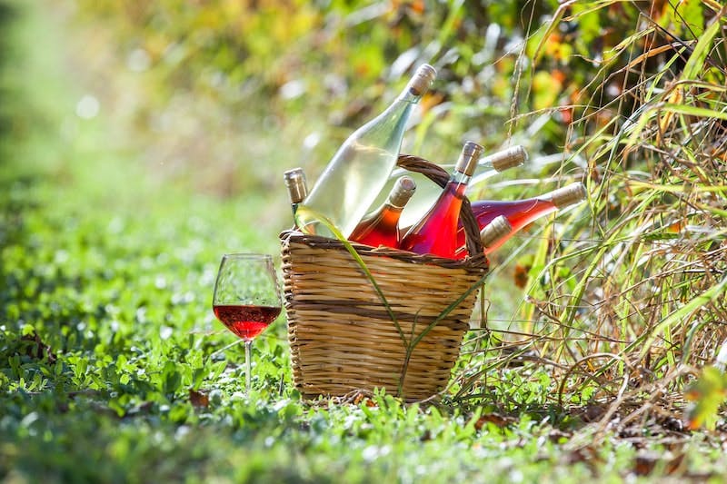 A wicker basket full of light red wines, and a glass alongside in the grass. 