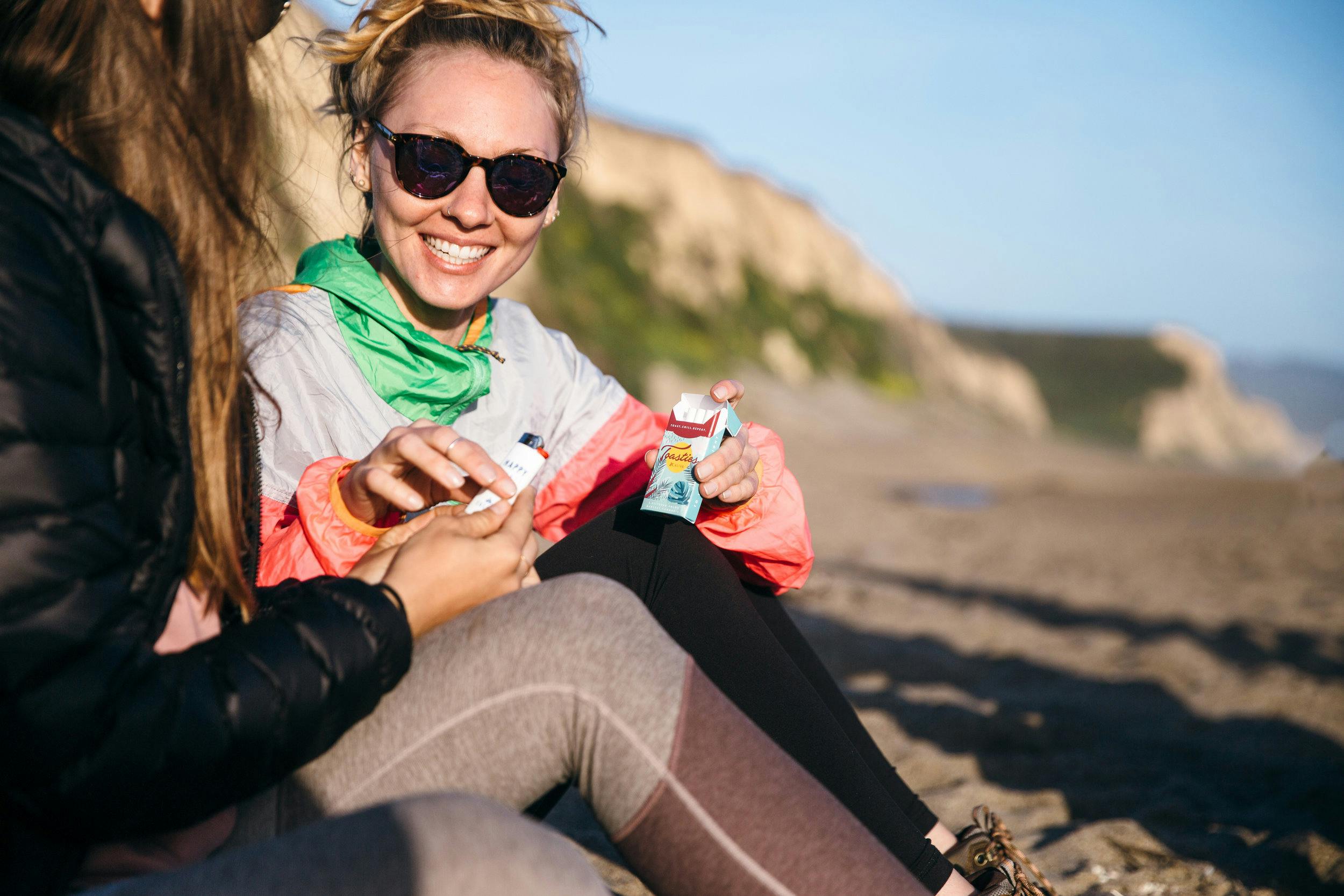 Two women sitting at the beach together sharing a Caliva Toasties cannabis cigarette.