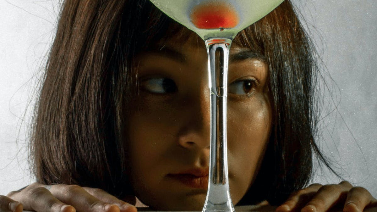 A woman stares blankly behind a glass with green liquid and a maraschino cherry, with her hands clinging to a tabletop.