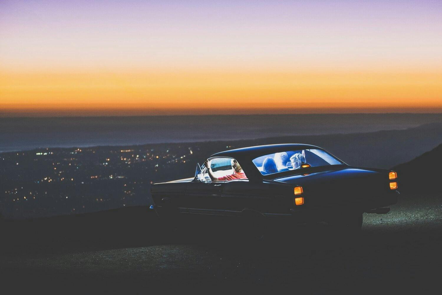 4 people sitting inside a car from a scenic point overlooking the city during sunset.