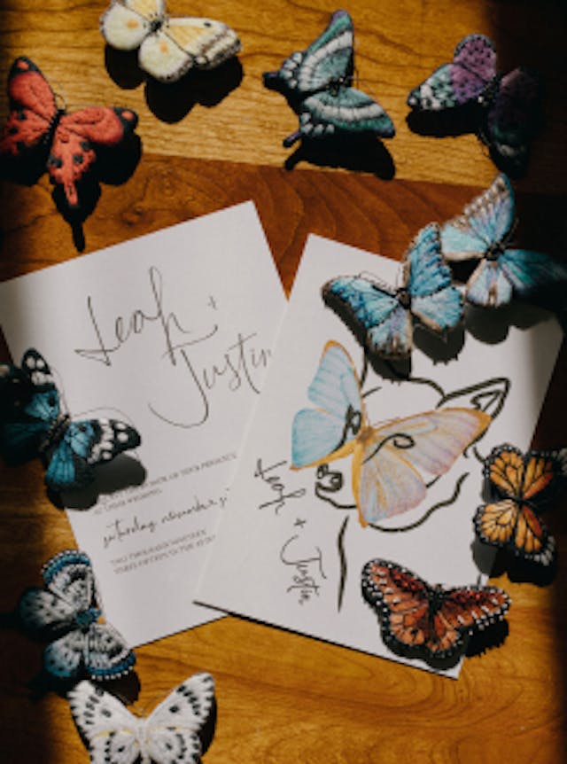 Decorative butterflies on hardwood floor with wedding invites that read "Leah and Justin" featuring an illustration of a dog with a butterfly mask. 