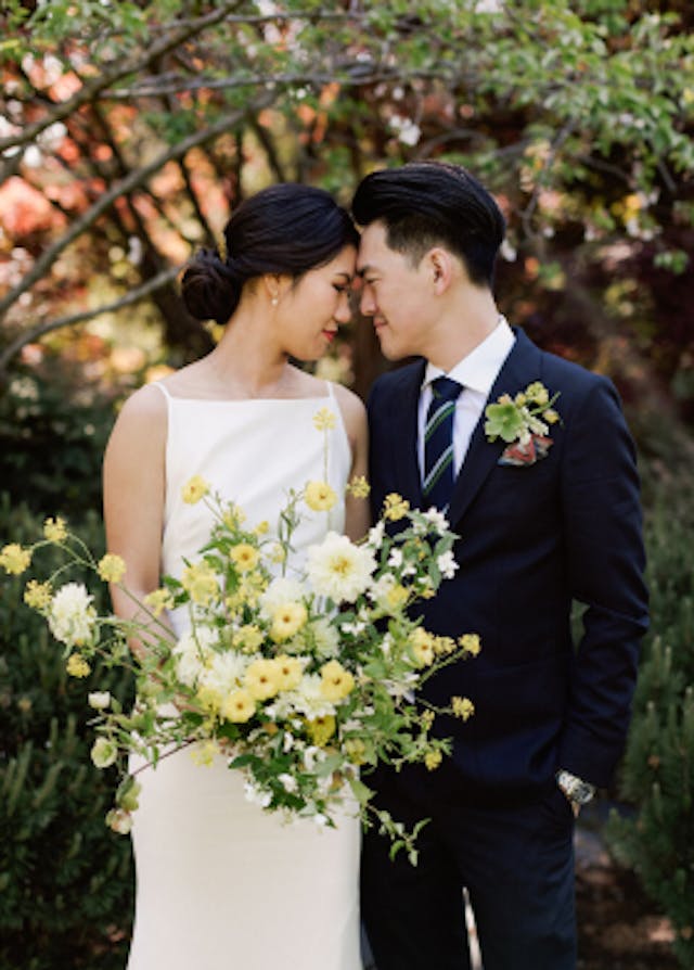 Bride holding white and yellow bouquet while touching foreheads with groom with trees in the background.