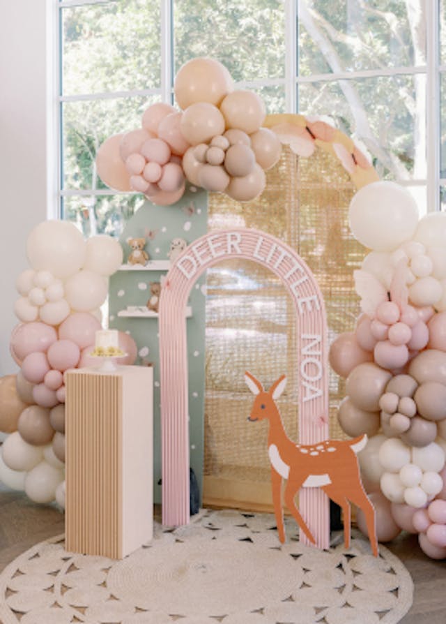 A deer-themed first birthday party with balloons, featuring an arch that reads "deer little noa" and a cake on a cake stand.