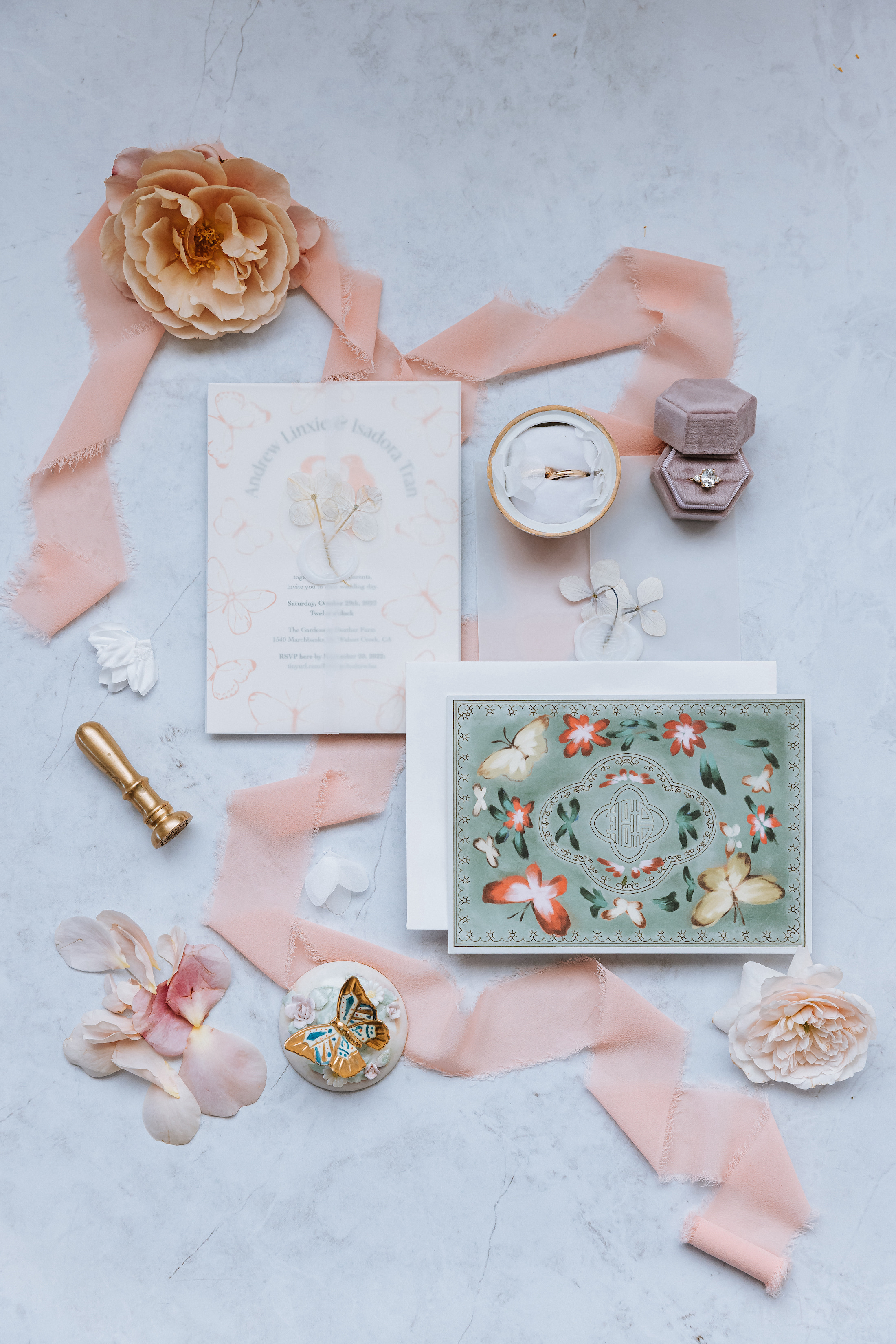 Flat lay featuring butterfly-themed wedding invitations surrounded by pink ribbon and wedding rings. 
