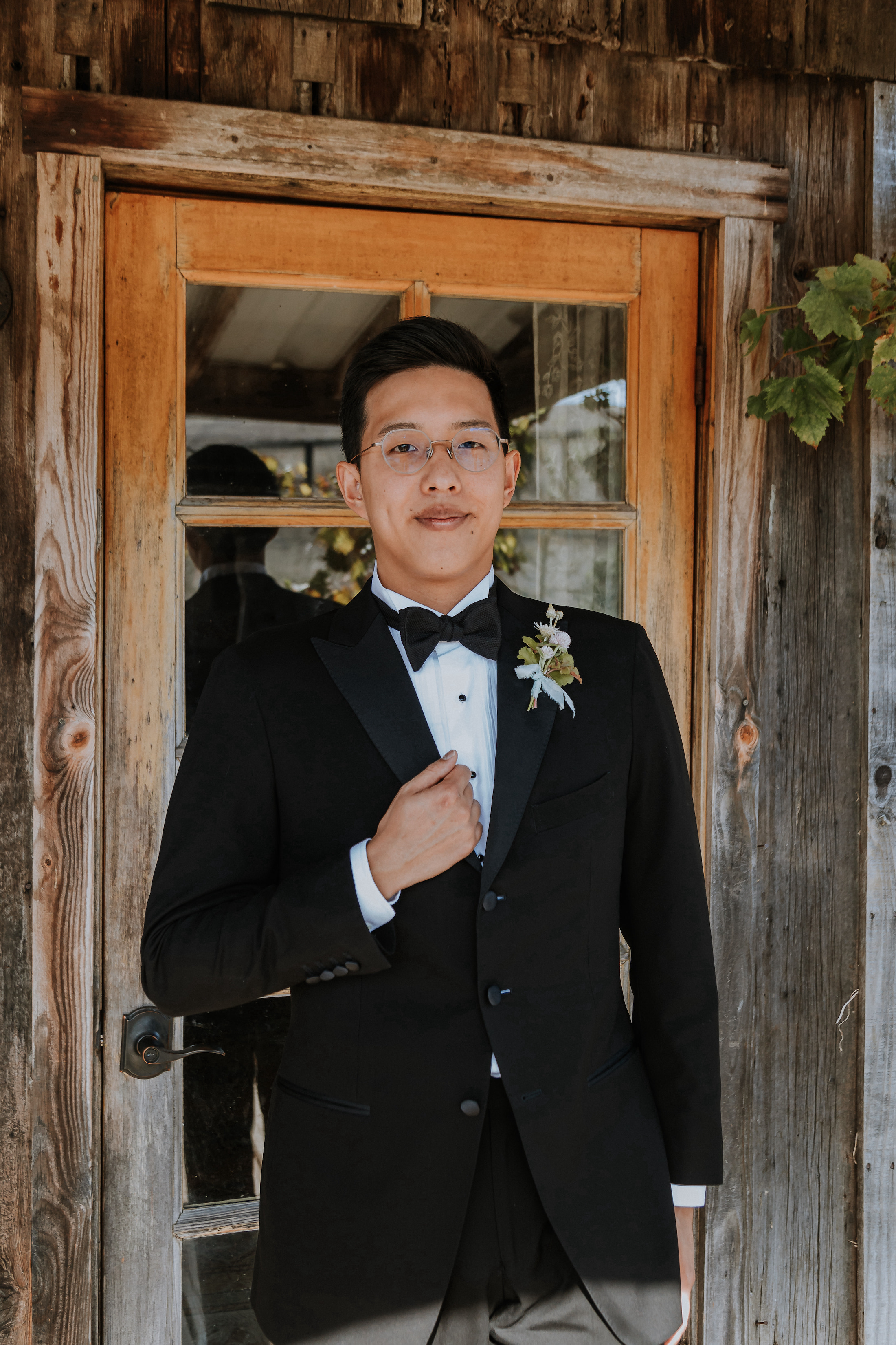 A groom in a tuxedo and bowtie smiling in front of a wooden barn.