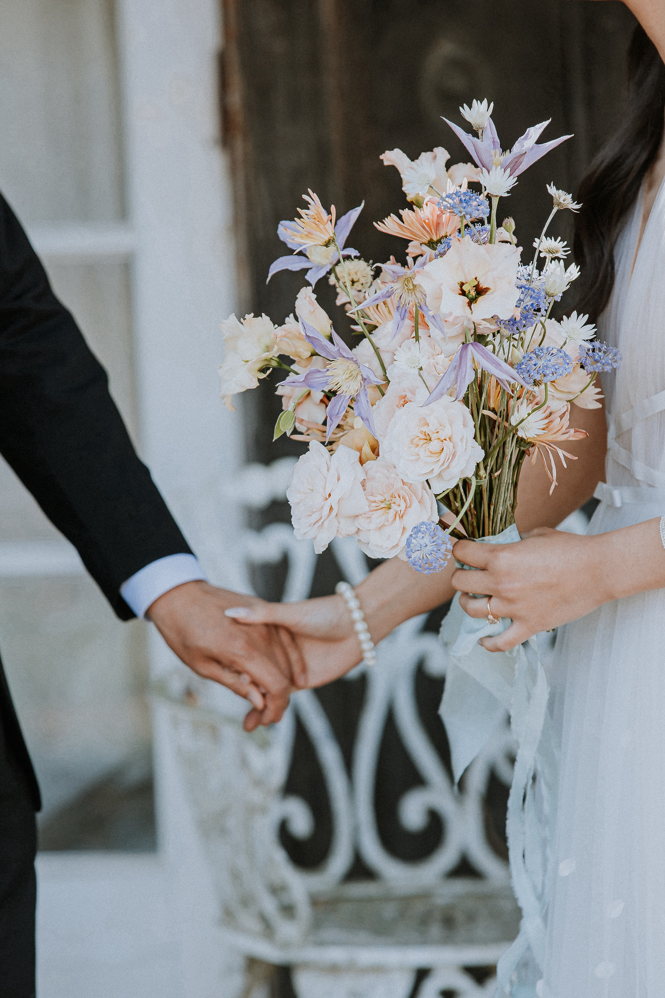 Close up of a bride and groom holding hands while the bride holds a pastel purple and peach colored bouquet.