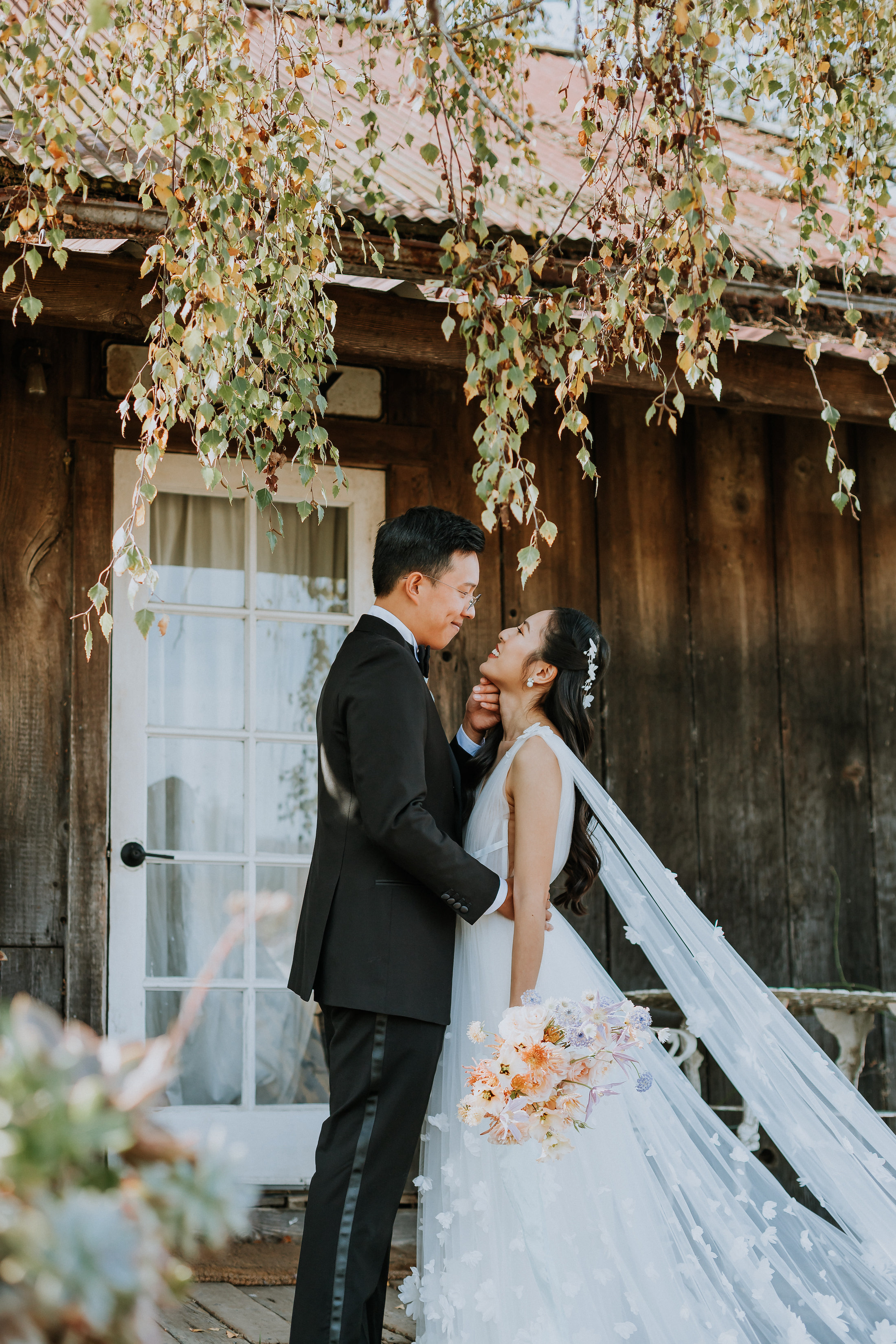Bride and groom face-to-face smiling at each other with the groom's hand on the bride's chin in front of rustic barn. Bride is holding bouquet down at her side.