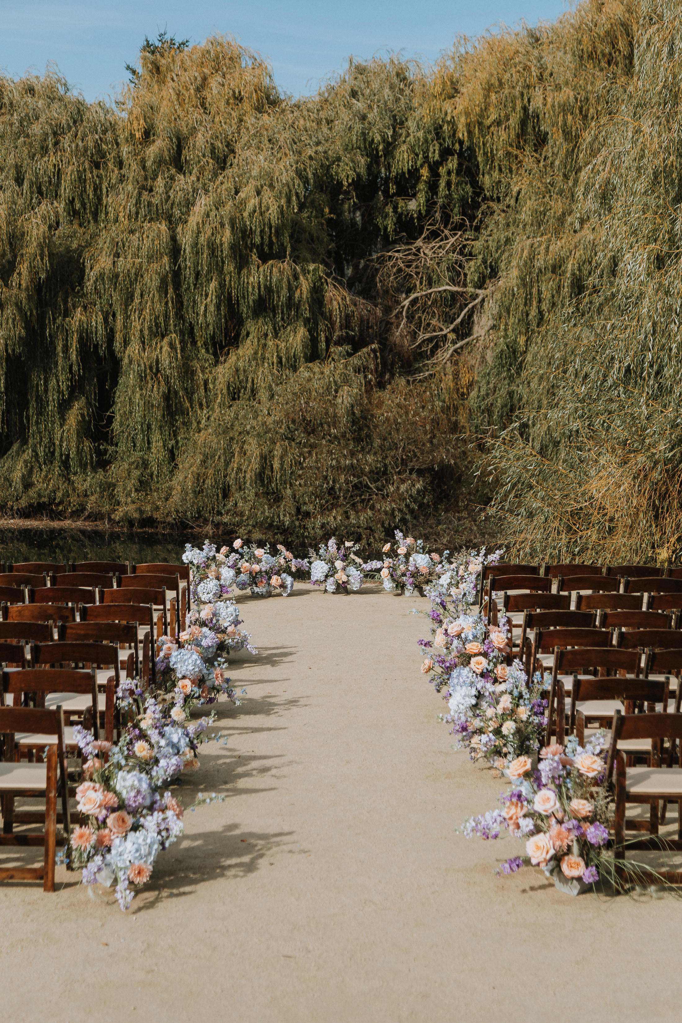 Lakeside ceremony aisle lined with peach, blue, and purple floral arrangements. 