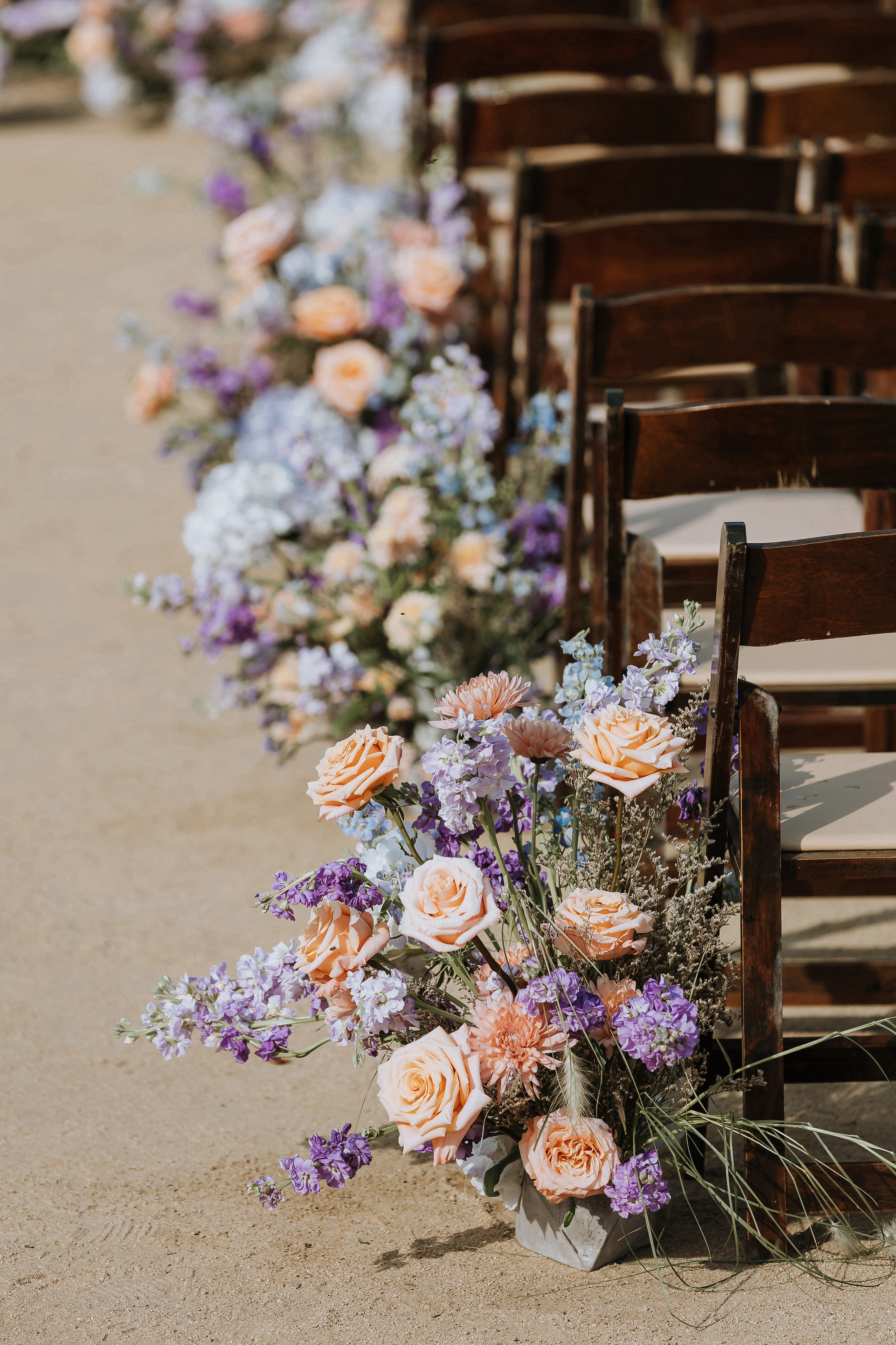 Close up of peach, blue, and purple floral aisle arrangements with dark brown chairs.