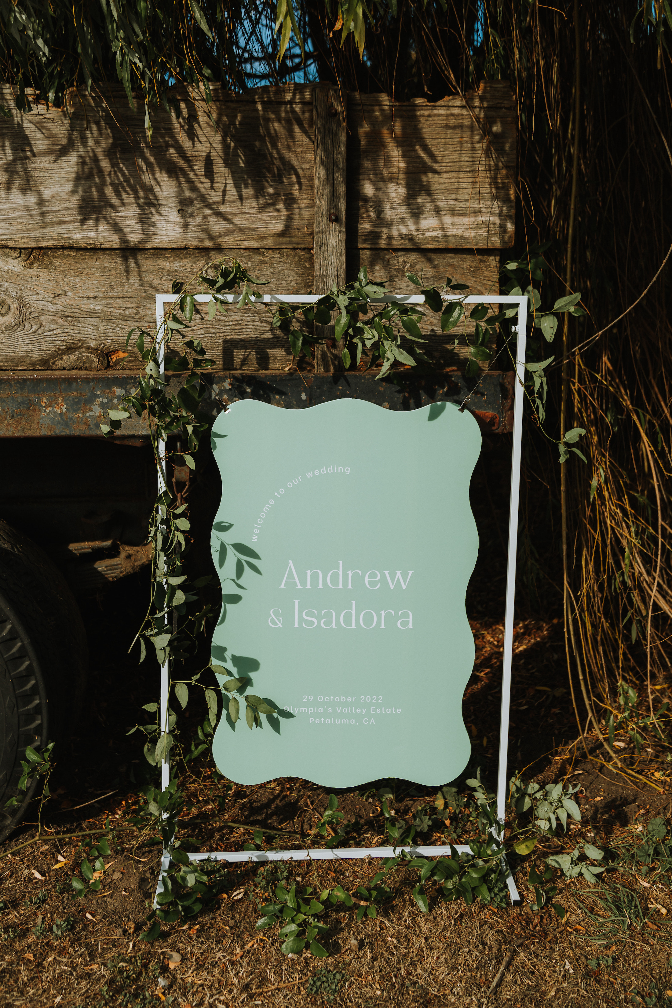 Sea foam colored hanging welcome sign with squiggle edges that reads "Welcome to our wedding. Andrew & Isadora. 29 October, 2022. Olympia's Valley Estate Petaluma, CA" 